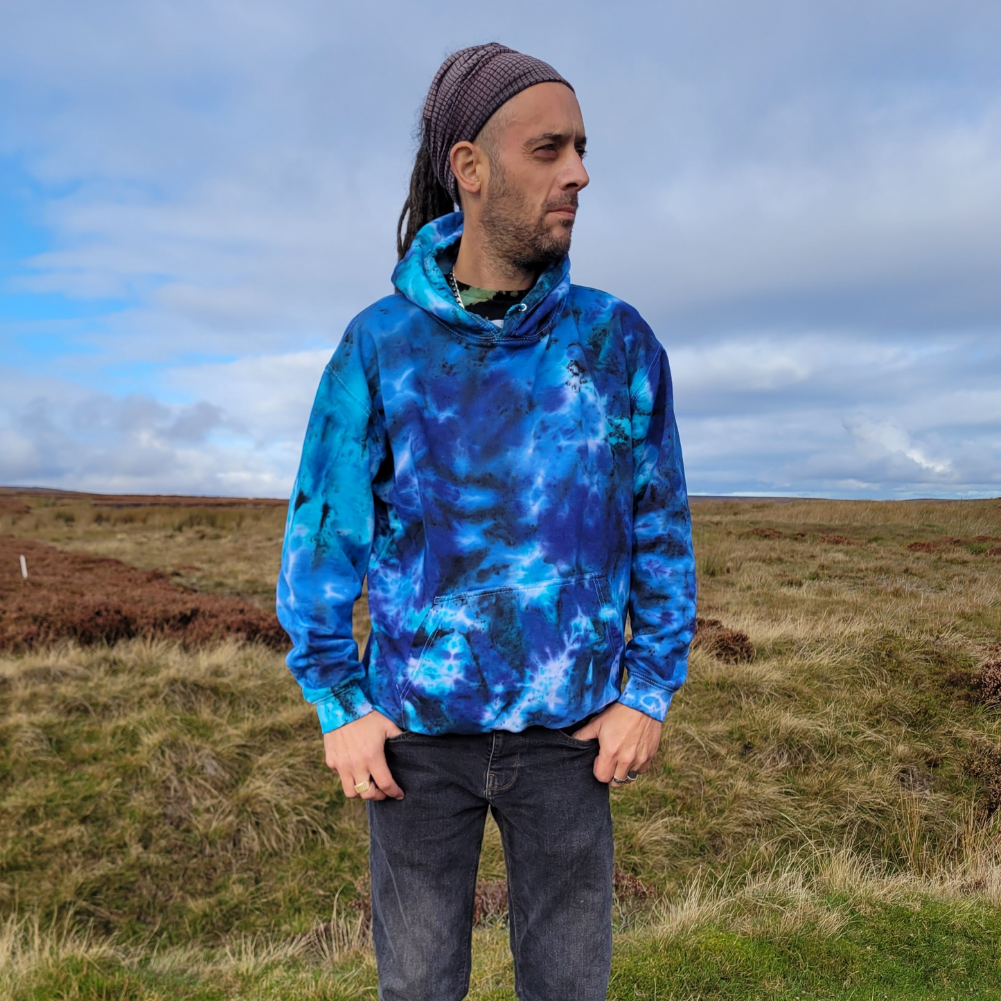 Person wearing a blue tie-dye hoodie standing in a grassy field with a cloudy sky.