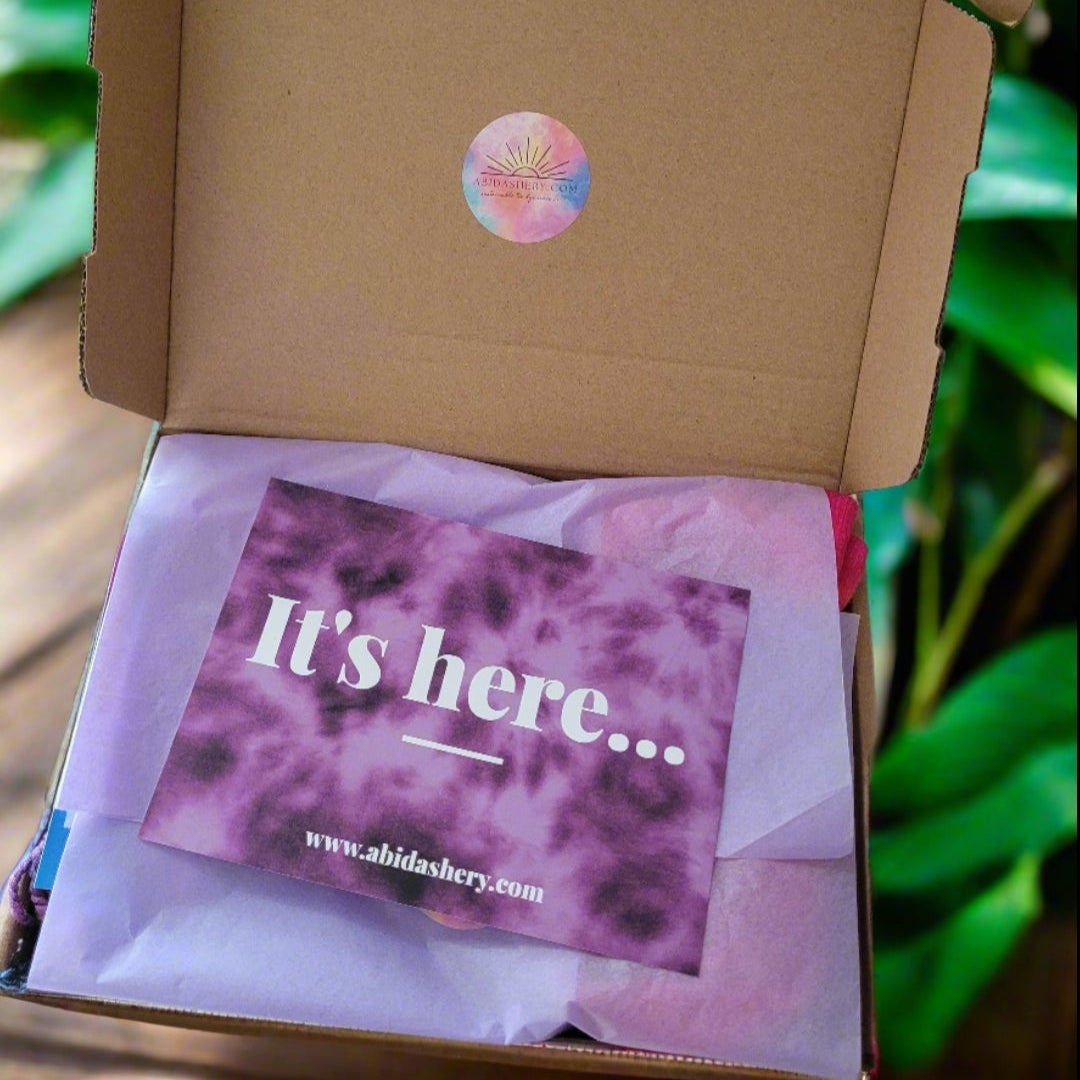 A mystery box with a pink and white label that says 'It's here...' and a green plant in the background.